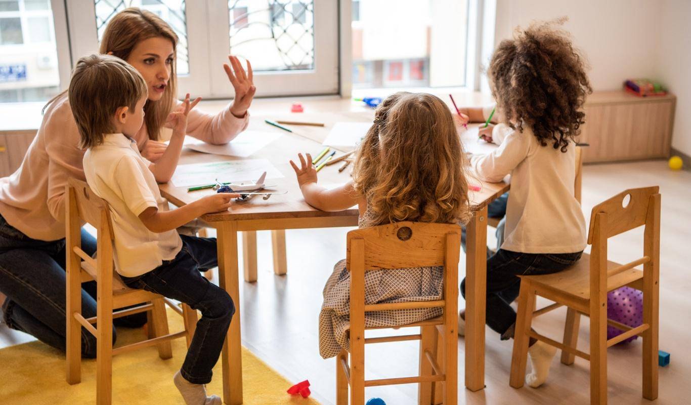 Professora branca reunida com três crianças em uma sala de aula infantil