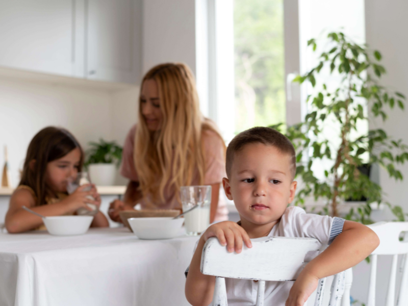 Foto de um bebê branco em uma cadeira olhando triste para o lado, com a mãe e uma irmã pequena ao fundo da foto