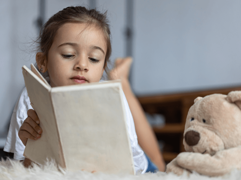 Foto de uma menina lendo um livro ao lado de um ursinho de pelúcia