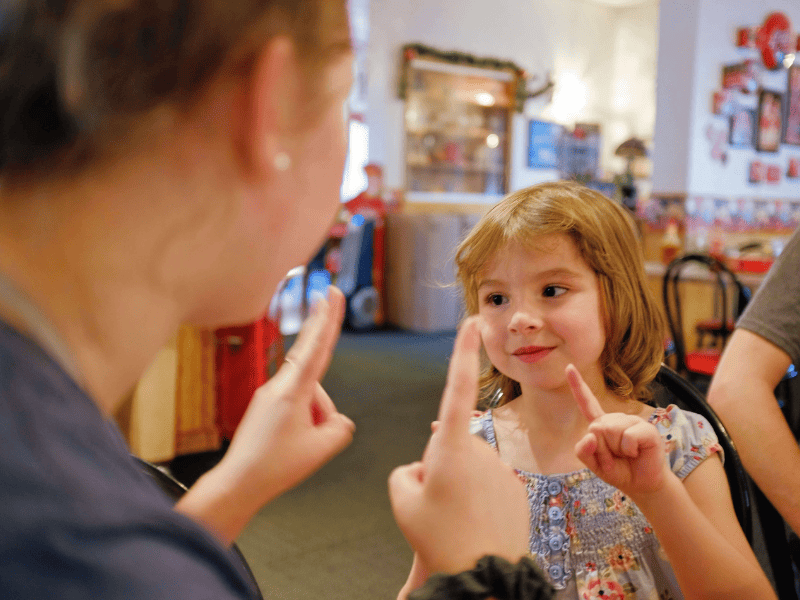 Mulher branca com as mãos levantadas, brinca com menina loira
